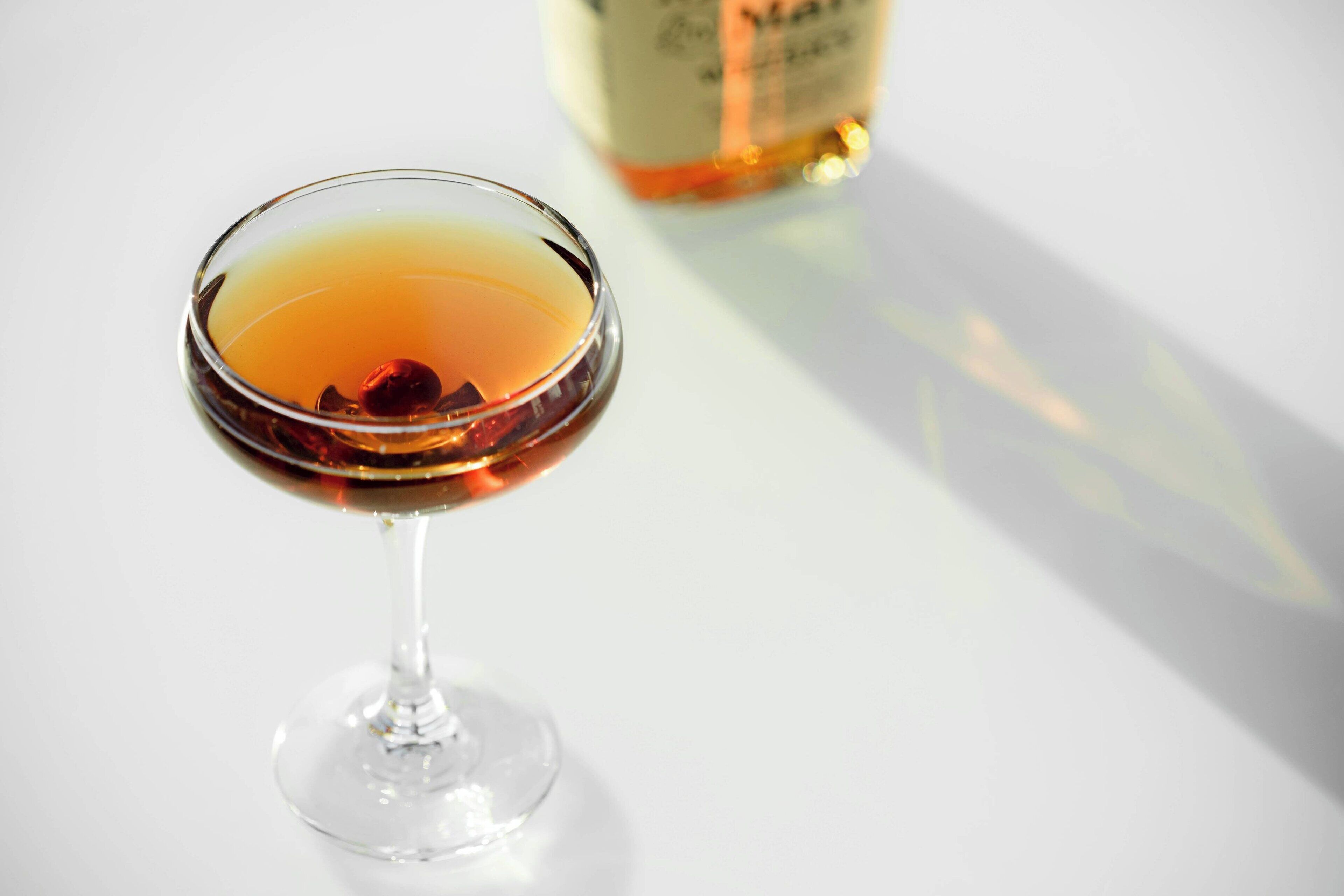 A cocktail in a stemmed glass with a blurred bottle in the background.