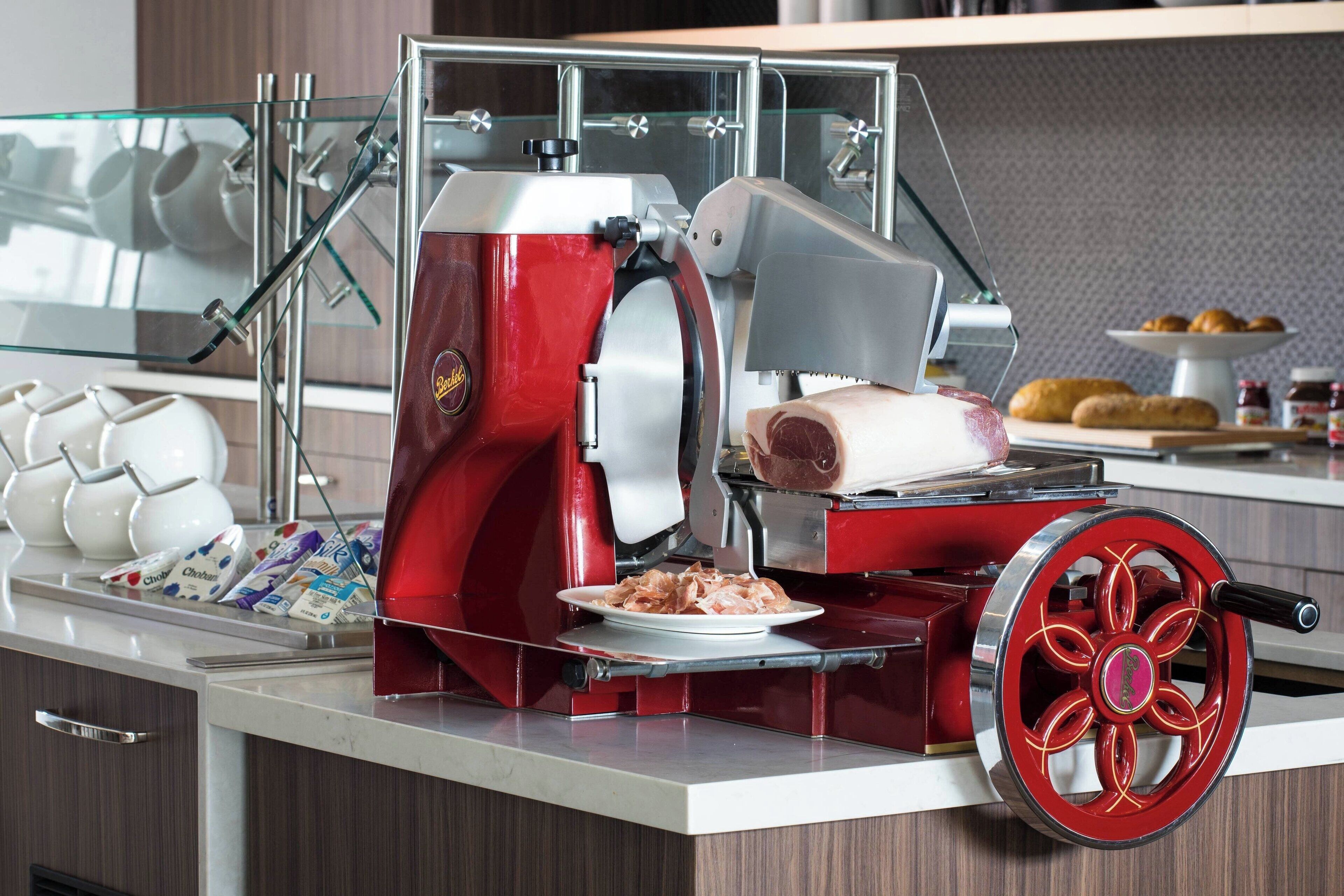 Une cuisine équipée d’une trancheuse à viande rouge, d’une vitrine en verre contenant des théières blanches et d’une assiette de viande tranchée.