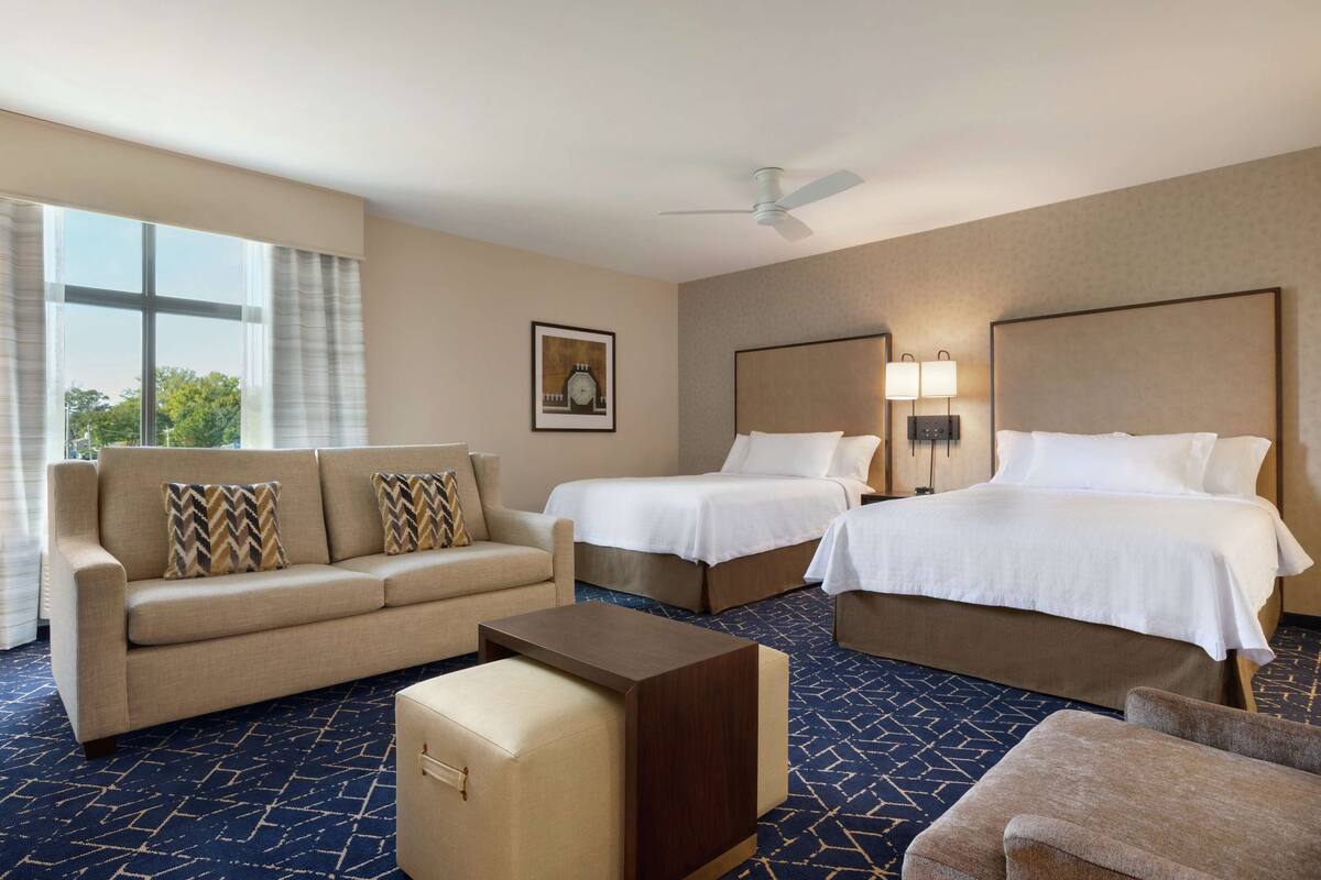 Een hotelkamer met een bank, twee bedden, een bijzettafeltje en een groot raam met gordijnen.