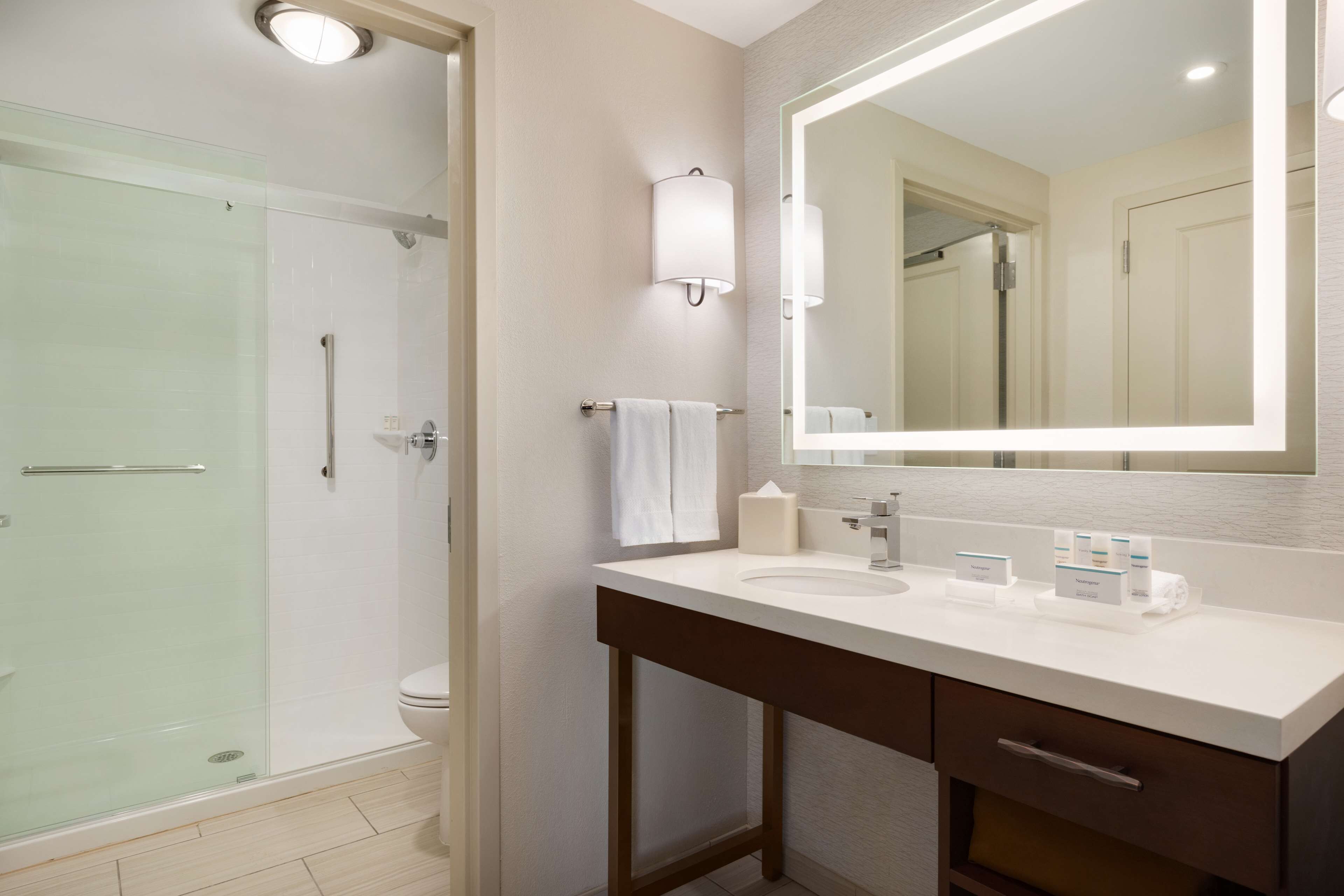 Une salle de bain moderne avec une douche à parois de verre, un grand miroir et un lavabo blanc agrémenté d’articles de toilette.
