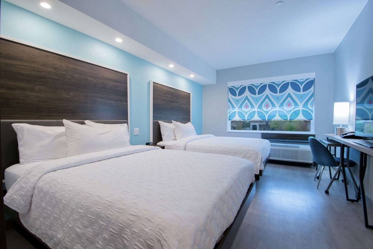 A hotel room with two beds, a desk, a chair, and a window with a geometric pattern.