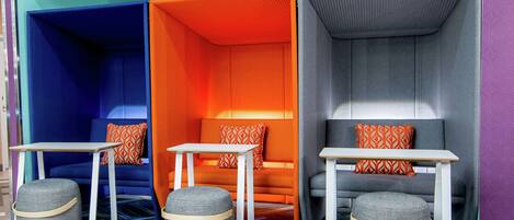 A hotel room with a vibrant color scheme, featuring orange, blue, and grey tones. It includes a sofa, two tables, and two ottomans.