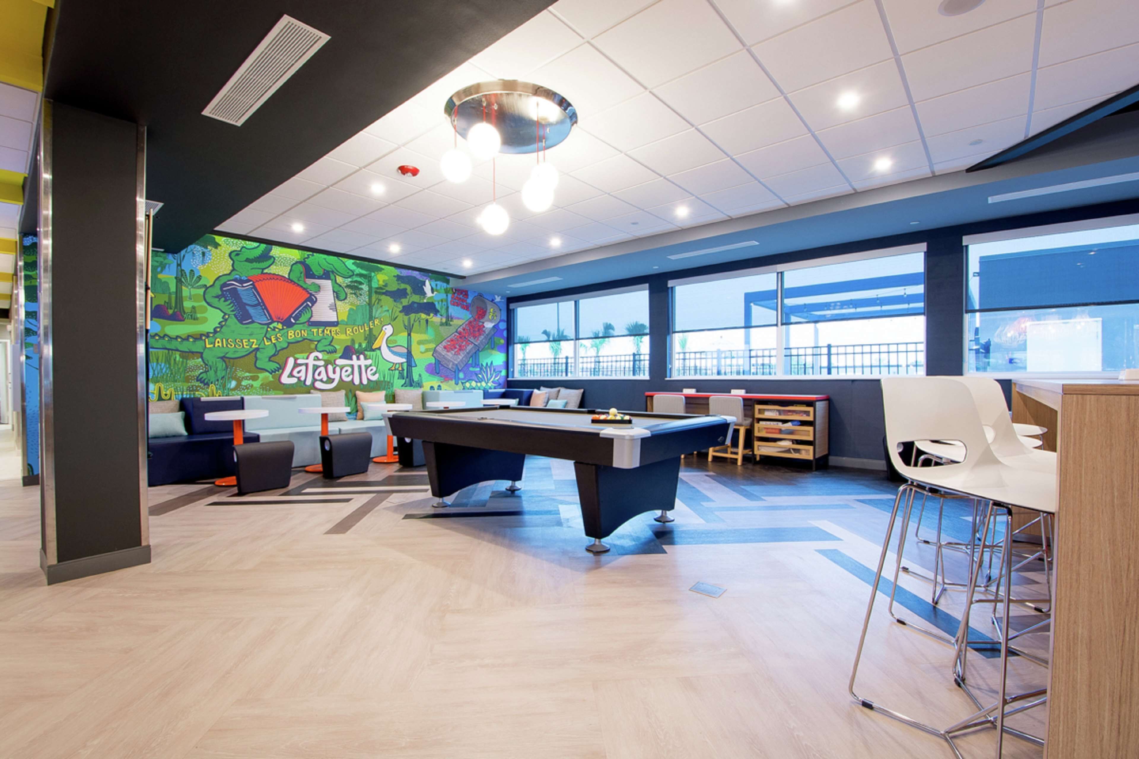 A pool table in a brightly lit room with a mural on the wall, large windows, and modern furniture.