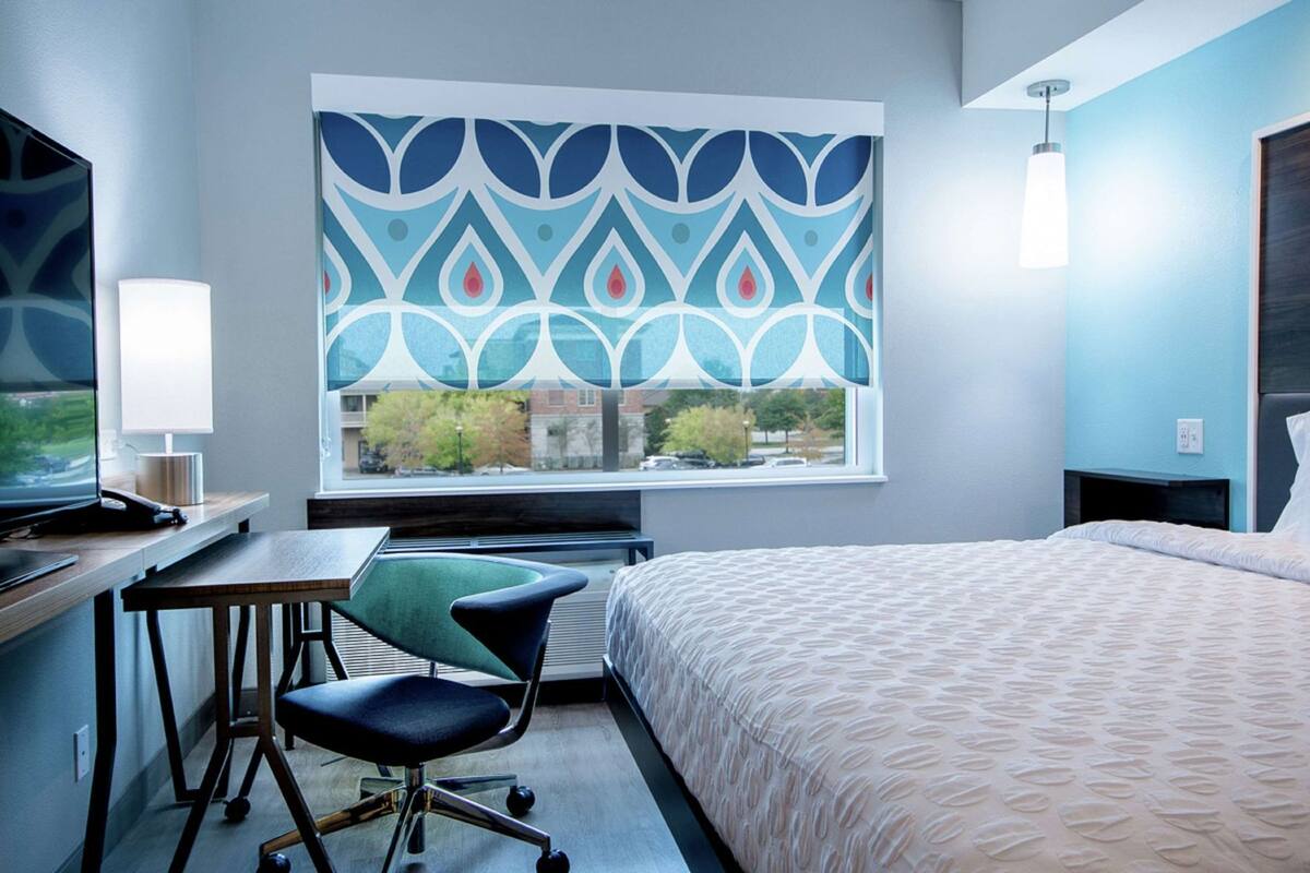 A hotel room with a bed, desk, chair, and a window with a patterned view.