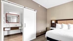 A hotel room with a bed, a nightstand with a lamp, a wardrobe, and a bathroom with a mirror and sink. - Hampton Inn & Suites Logan (Logan)