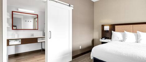 A hotel room with a bed, a nightstand with a lamp, a wardrobe, and a bathroom with a mirror and sink.