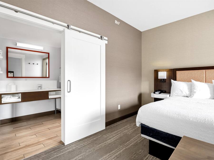 Una habitación de hotel con una cama, una mesita de noche con lámpara, un armario y un baño con espejo y lavamanos.