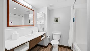 A modern bathroom with a white sink, mirror, and toilet. There are towels stacked on a shelf and a shower curtain.