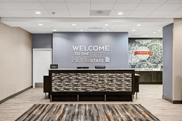 A reception area with a 'Welcome to the Bee Hive State' sign, a modern counter, and a patterned rug. - Hampton Inn & Suites Logan (Logan)