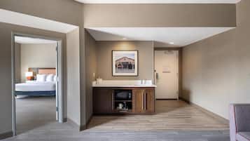 A hotel room with a wooden floor, a bed, a bathroom with a sink and mirror, and a hallway leading to another room.