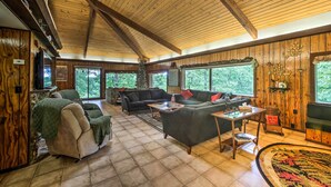Uma sala de estar espaçosa com paredes de madeira e teto alto, com lareira, diversas opções de assento e janelas grandes que oferecem vista para o exterior.