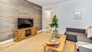 A modern living room with a wooden wall panel, a flat-screen TV on a wooden entertainment unit, a grey sofa, a coffee table with decorative items, and a potted plant.
