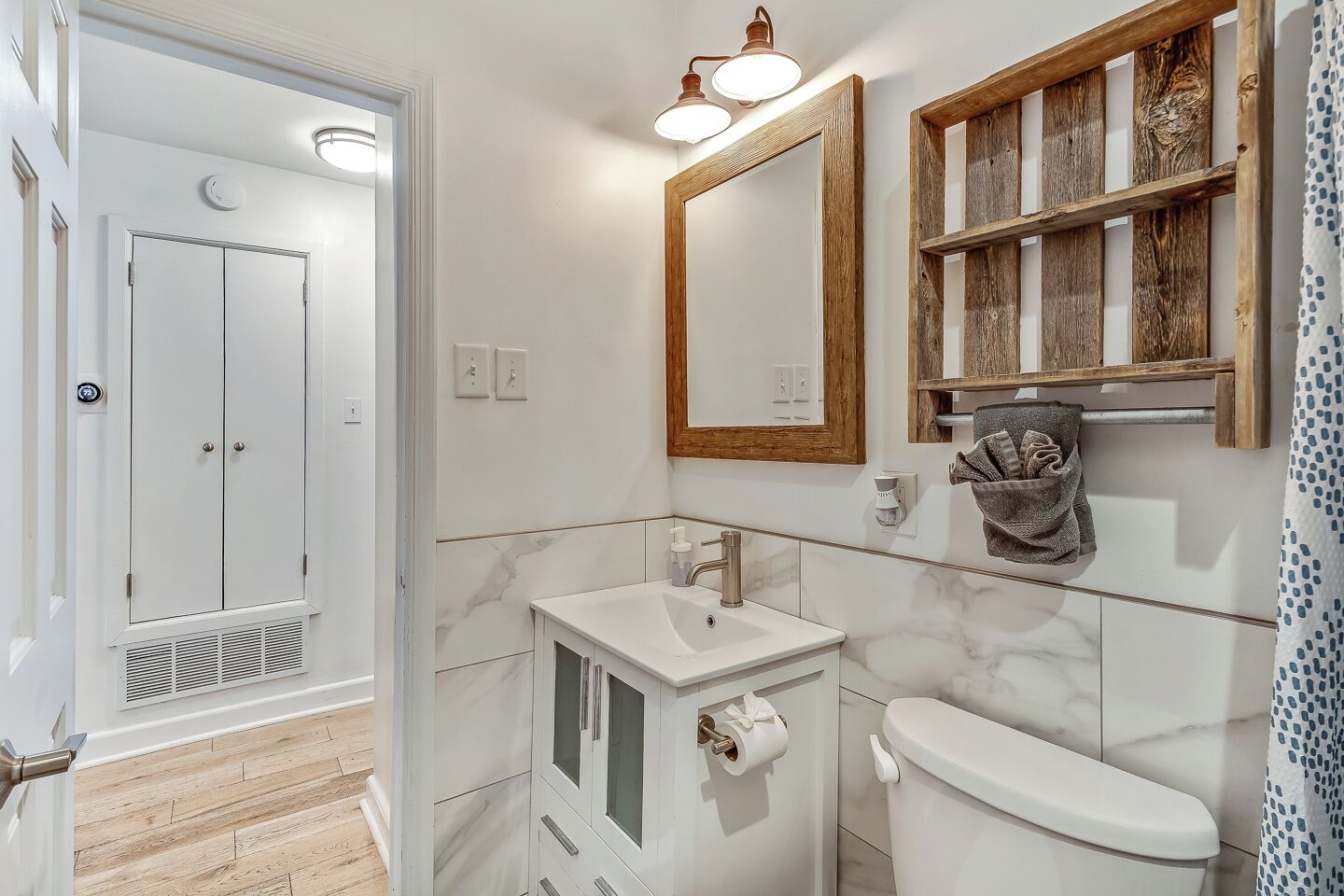 Baño con lavamanos blanco, inodoro y espejo con marco de madera. Se observa un perchero con una toalla y cortina de ducha estampada con lunares.