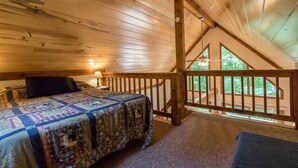 Een slaapkamer met een houten plafond, een bed met een sprei, een nachtkastje en uitzicht op de omgeving via een balkon.