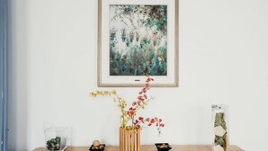 Ein Holztisch mit einem gerahmten abstrakten Gemälde, einer Holzvase mit Blumen und zwei Glasbehältern mit Dekoration.