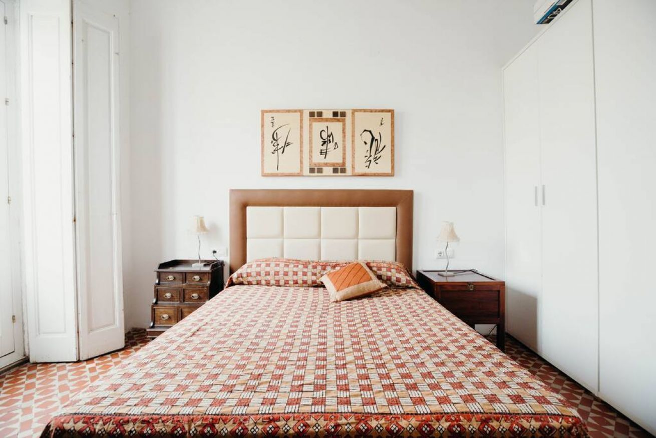 A bedroom with a patterned bedspread, a headboard, two bedside tables, a lamp, and a wall with framed calligraphy.