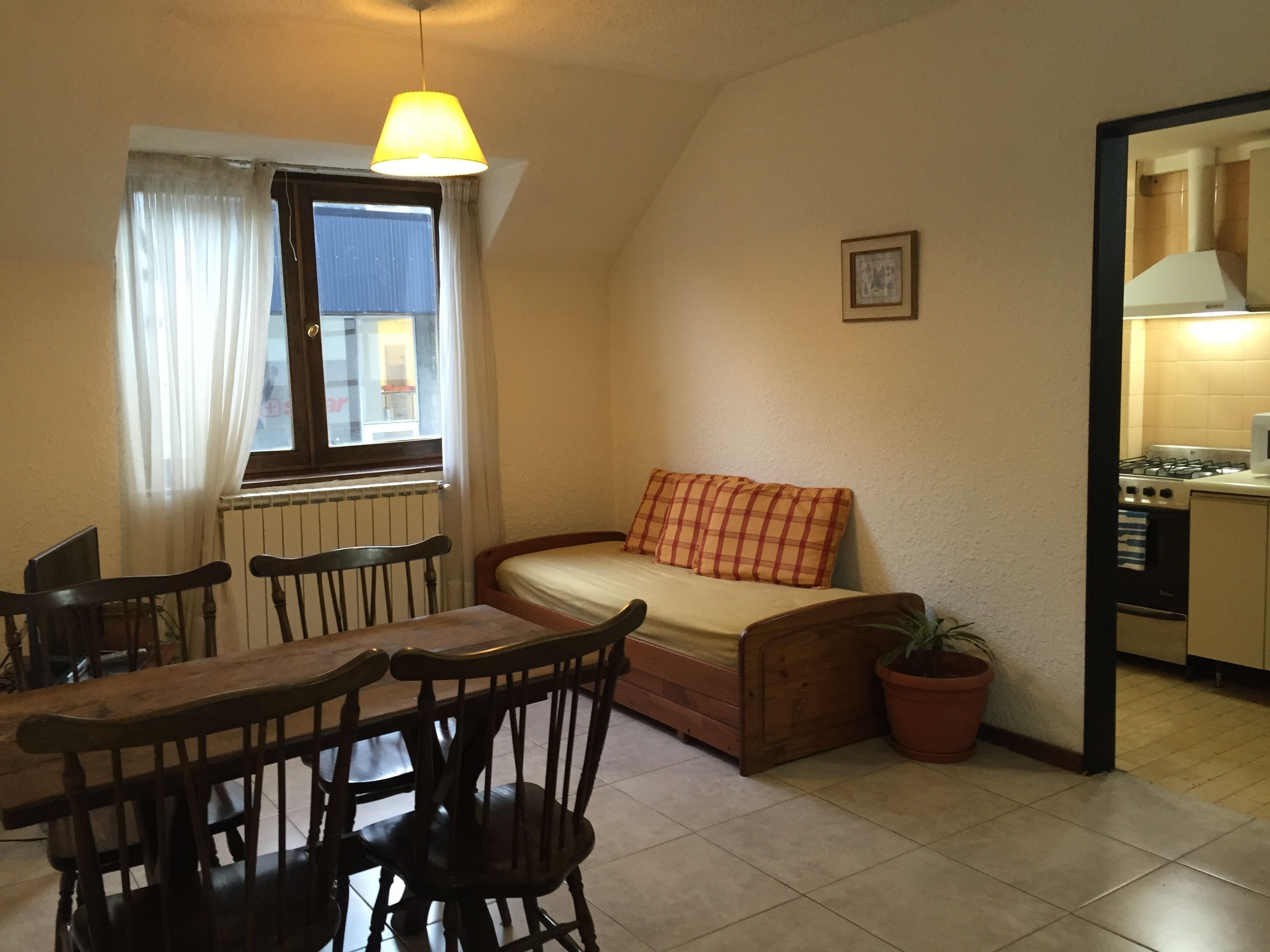 Un espacio habitable compacto con una mesa de comedor, sillas, un sofá, una planta en maceta, una ventana con cortinas y una kitchenette.