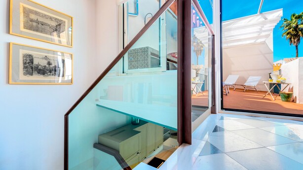 Een modern interieur met een glazen trap, afbeeldingen in lijsten aan de muur en uitzicht op het buitenterras met ligbedden.