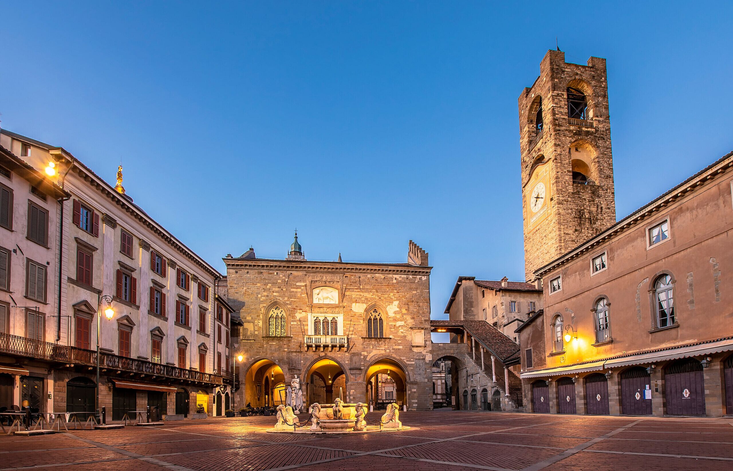 Una plaza histórica con una imponente torre del reloj, una fuente y arquitectura tradicional.