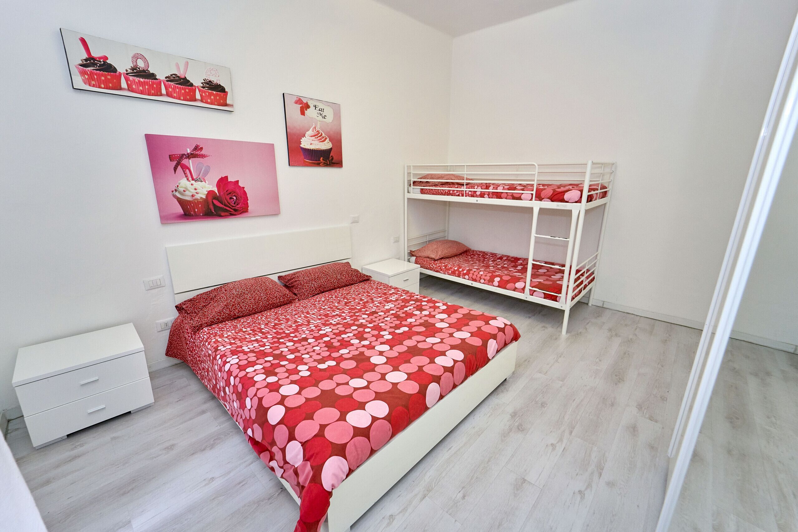 Ein Schlafzimmer mit Etagenbett, einem Einzelbett mit rotem Punktenmuster auf der Bettdecke, einem weißen Nachttisch und zwei Wandbildern mit Cupcakes.