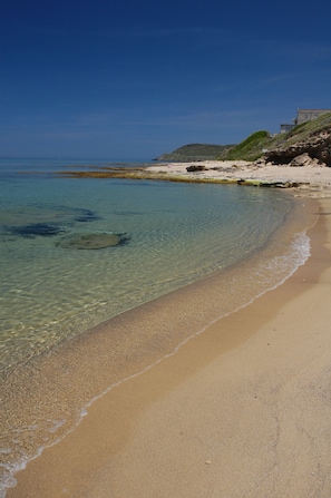 A beach with clear shallow waters, a sandy shore, and a building on a hill in the background.