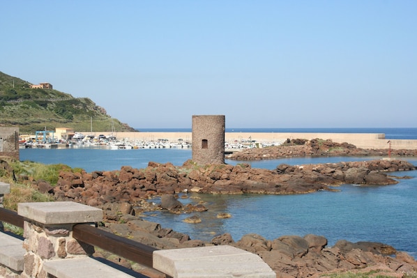 Paisaje costero con costa rocosa, una torre y un puerto deportivo con barcos.