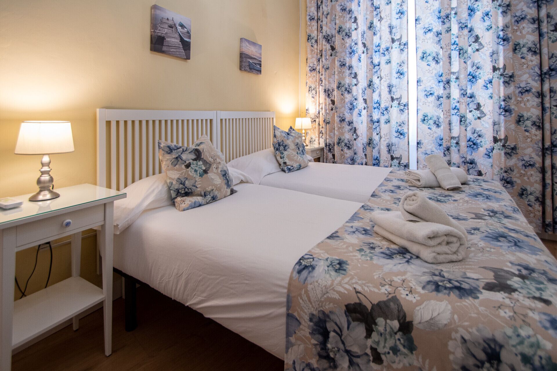 Une chambre d’hôtel avec deux lits, une table de chevet avec une lampe, des rideaux à motifs floraux et des œuvres d’art accrochées au mur.