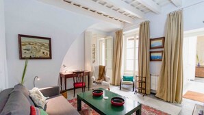 A living room with a sofa, a coffee table, a chair, and a desk. There are curtains on the windows, and a painting on the wall. - Duplex de la Botanica - Cadiz (Cádiz)