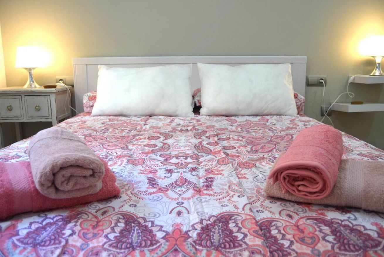 A bed with a patterned bedspread, two white pillows, and rolled towels on either side.
