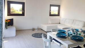 Un salon moderne avec un canapé blanc, une table à manger dressée avec de l’assiette bleue et deux fenêtres aux cadres sombres.