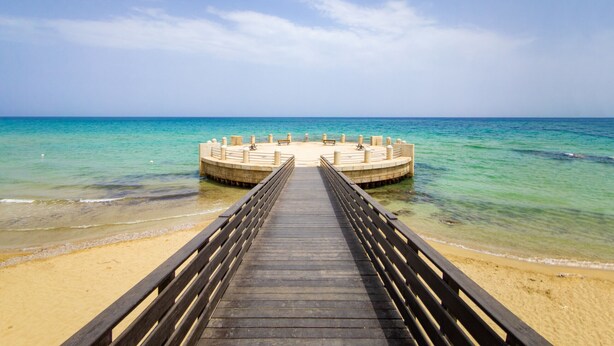 Une jetée en bois mène à une plateforme circulaire dotée de bancs, d’où l’on peut admirer une plage et une mer d’un bleu limpide.