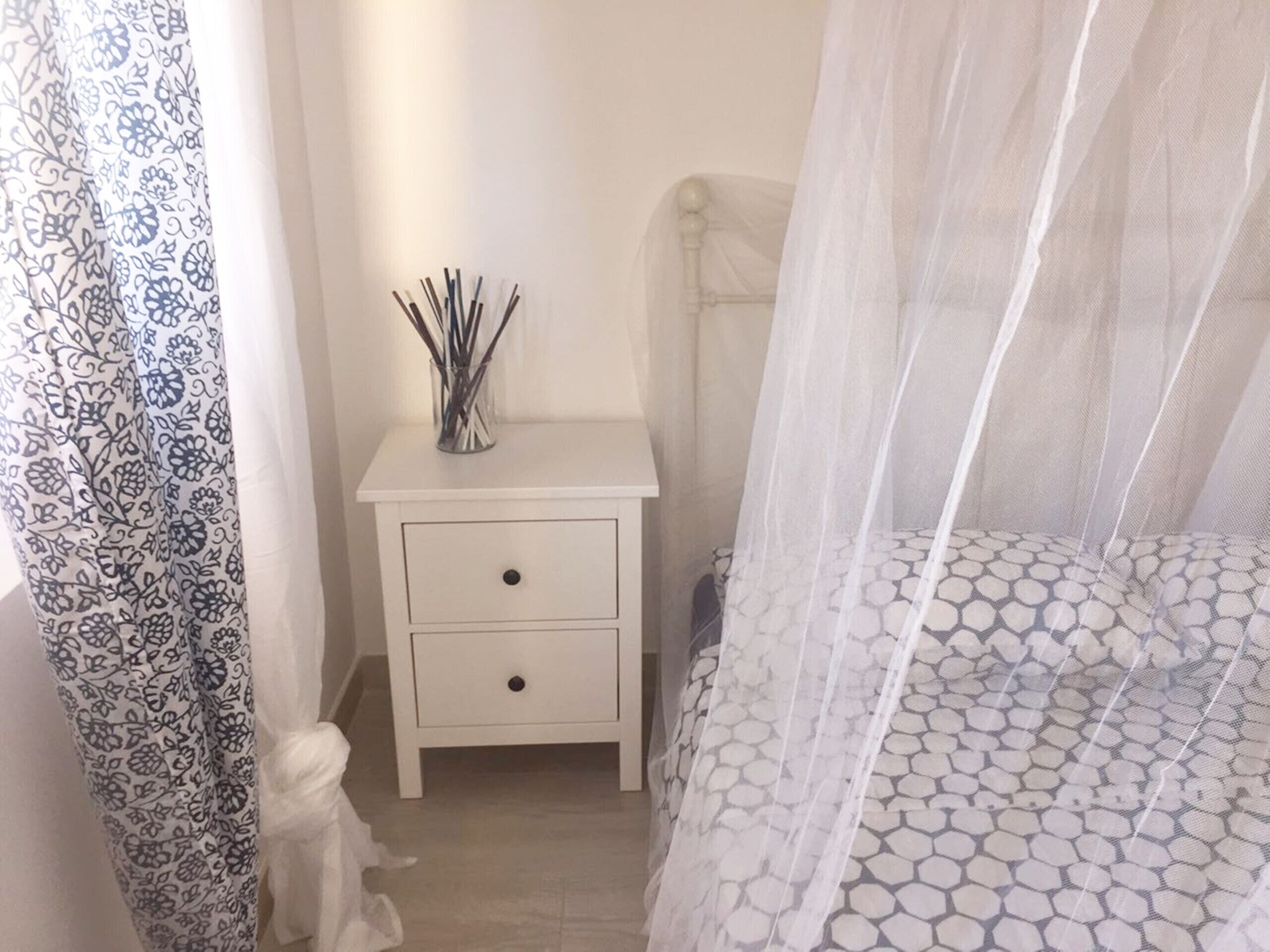 Ein Schlafzimmer mit einem weißen Nachttisch, einem Bett mit einem gemusterten Überwurf und einem Beistelltisch mit einer Vase voller Zweige, sowie einem durchsichtigen Baldachin über dem Bett.