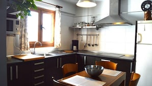 A compact kitchen with dark cabinetry, a wooden dining table, a hanging light fixture, and a window with curtains. - El Mirador de Cádiz (C��diz)