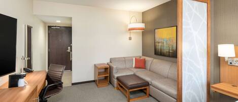 A hotel room with a wooden desk, a leather chair, a sofa, a small table, and a wall-mounted lamp.