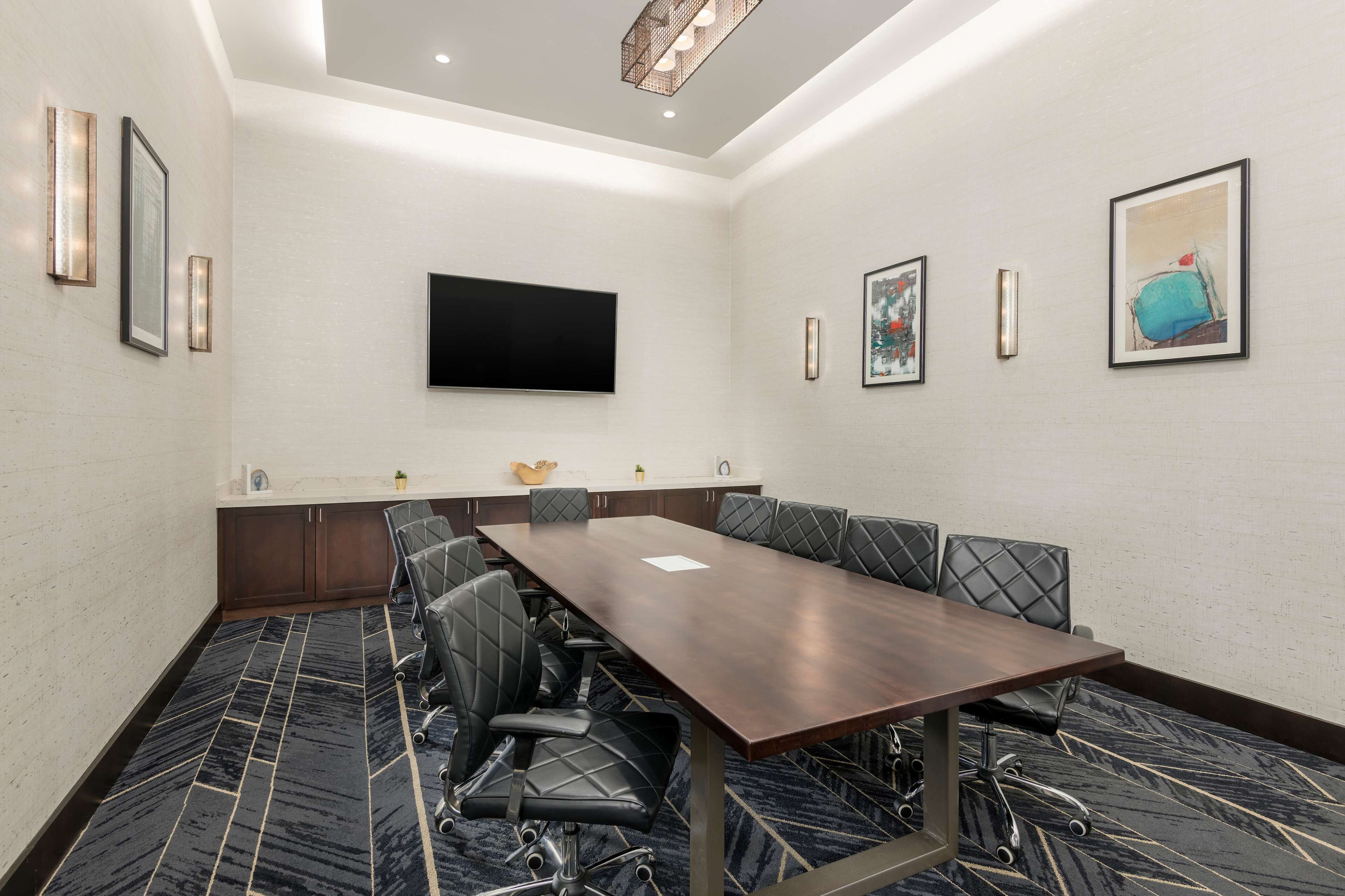A conference room with a long table, multiple chairs, a flat-screen TV, framed artwork, and a patterned carpet.