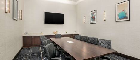 A conference room with a long table, multiple chairs, a flat-screen TV, framed artwork, and a patterned carpet.