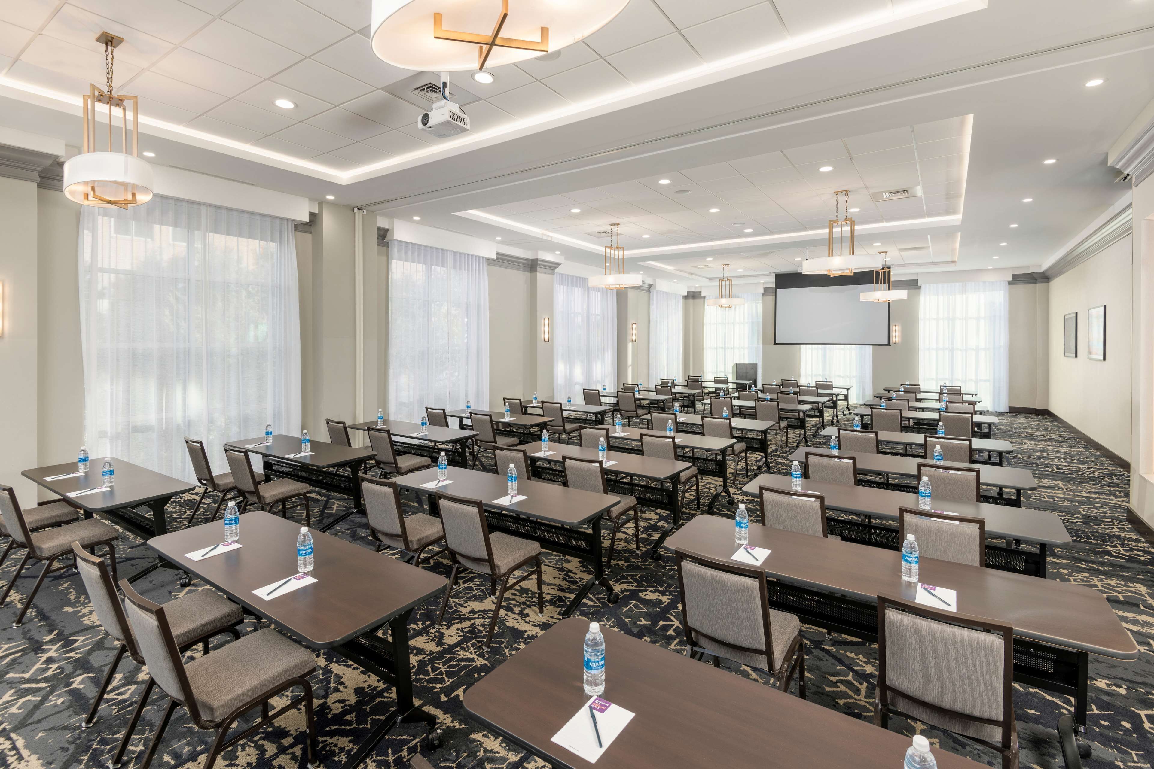 A conference room with multiple tables, chairs, and a large screen. Water bottles are placed on each table.