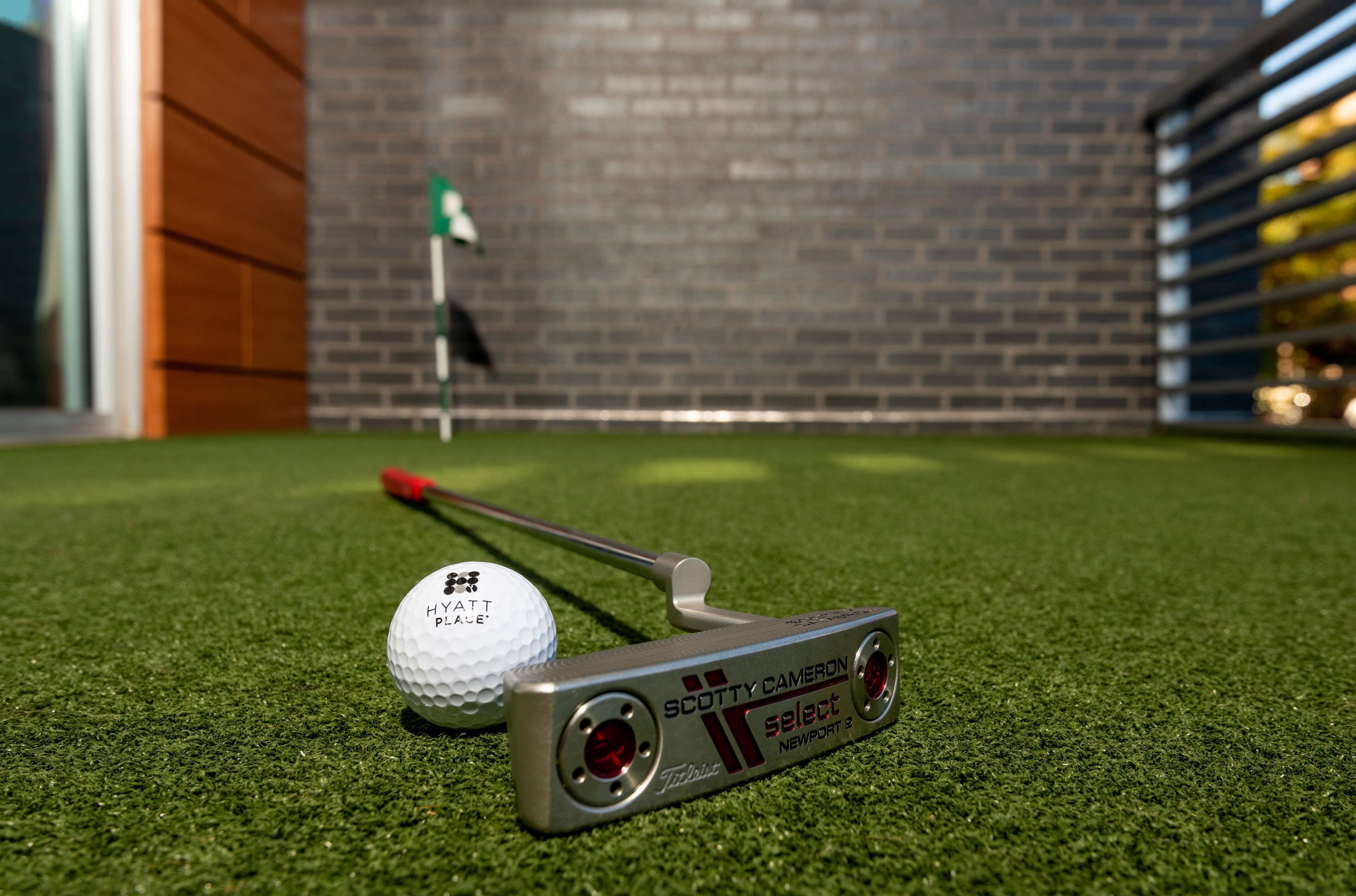 A golf ball and putter on artificial turf with a brick wall and a flag in the background.