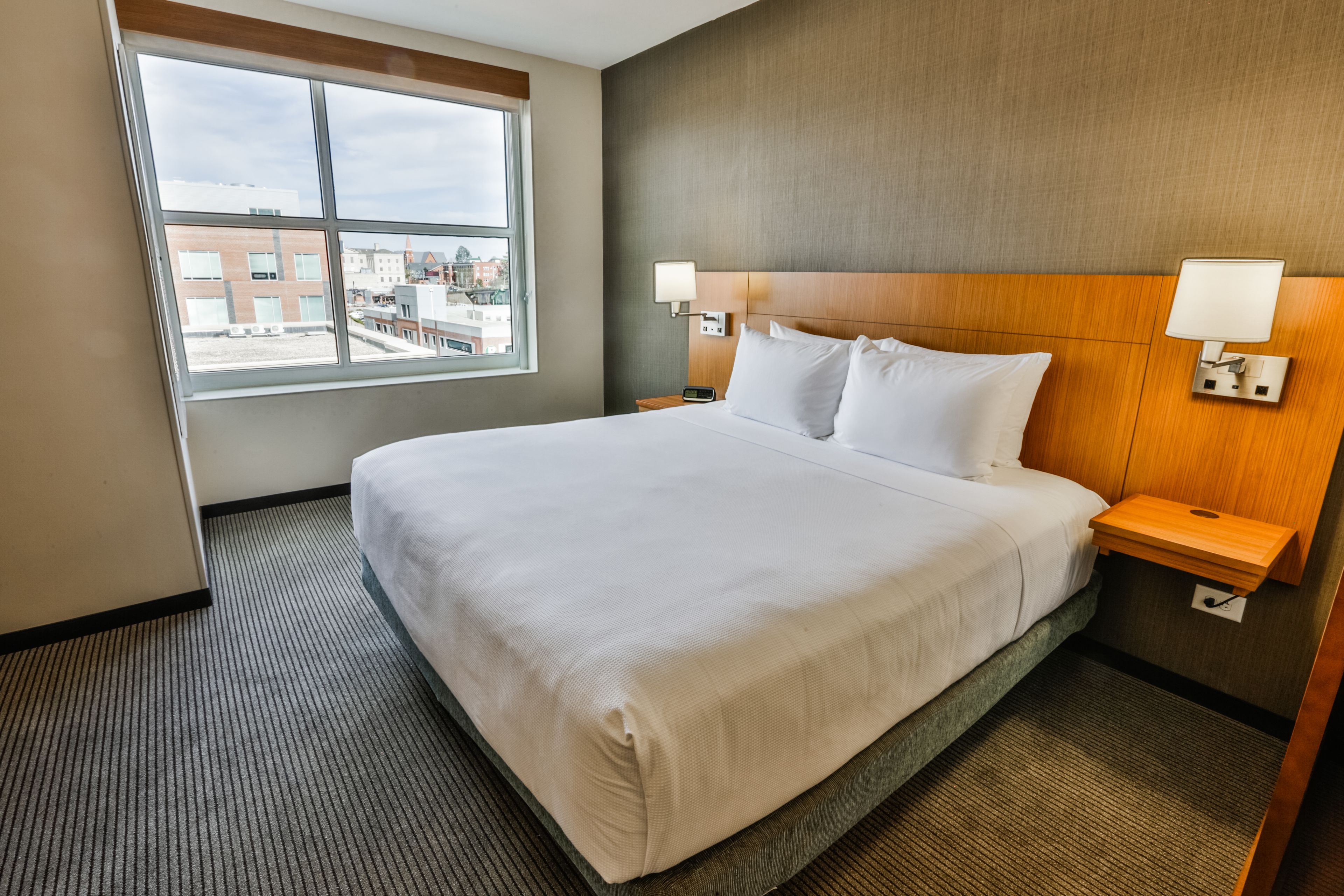 A hotel room with a large bed, two bedside tables, a window with a view of buildings, and a headboard with wall-mounted lamps.