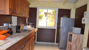 A compact kitchen with wooden cabinets, a built-in oven, a refrigerator, and a window overlooking a garden. - Escape with the sound of the waves (Deshaies)