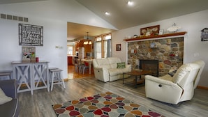 A spacious living room with a stone fireplace, comfortable seating, a dining area, and a kitchen visible through an open door. - Riesling Rod and Reel: Fishers Paradise (Honeoye)