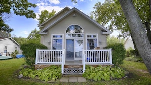 A two-story house with a front porch, a garage, and a small garden with green plants. - Riesling Rod and Reel: Fishers Paradise (Honeoye)