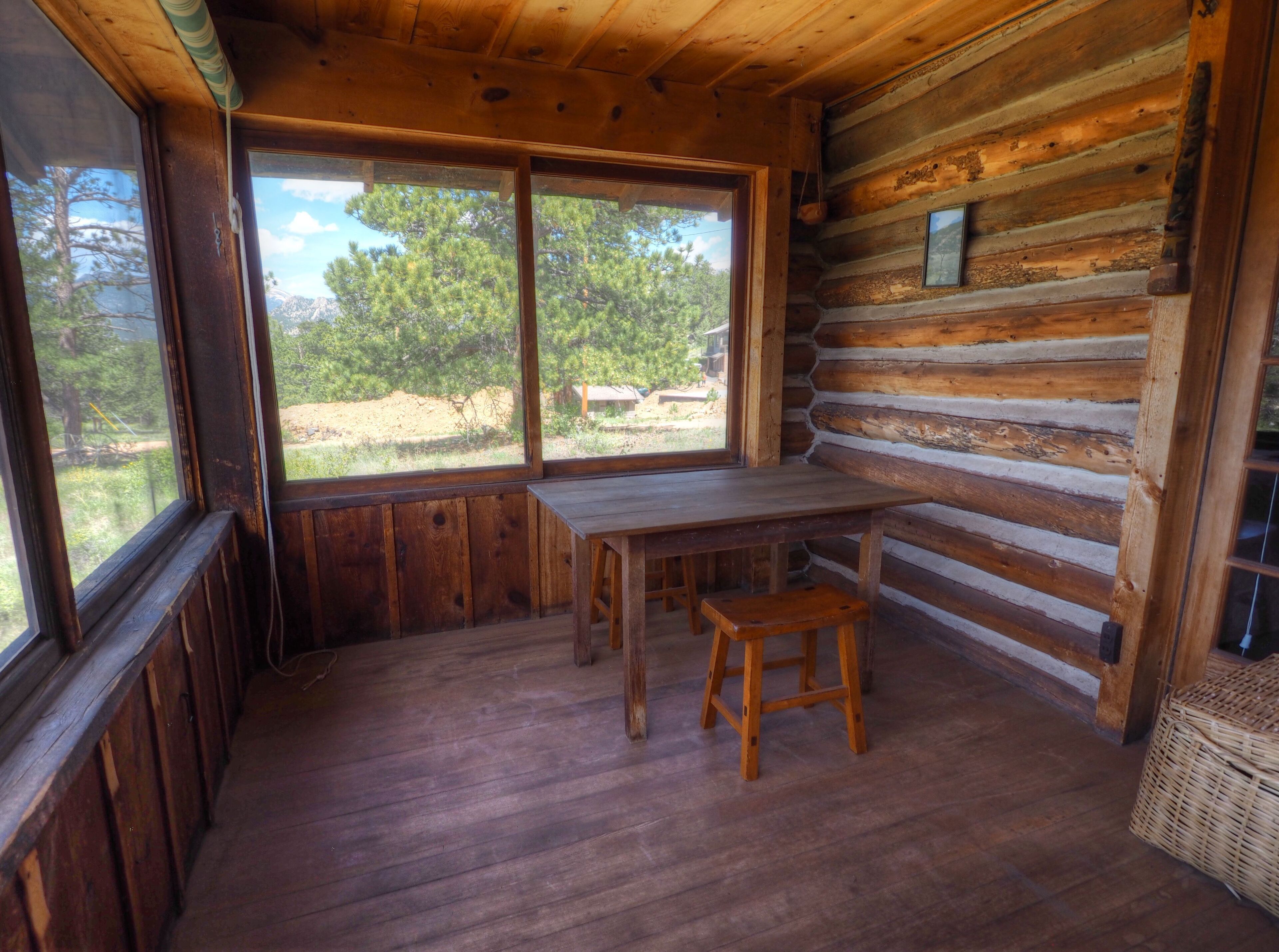 Interior de una cabaña de troncos con una mesa y banco de madera, ventanas grandes y vistas al exterior.