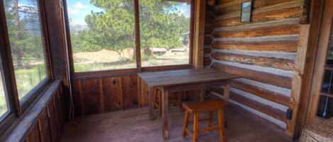 Interior de una cabaña de troncos con una mesa y banco de madera, ventanas grandes y vistas al exterior.