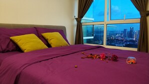 Una cama con ropa de cama morada y almohadas amarillas, un jarrón con flores, y vista a la ciudad a través de amplias ventanas.