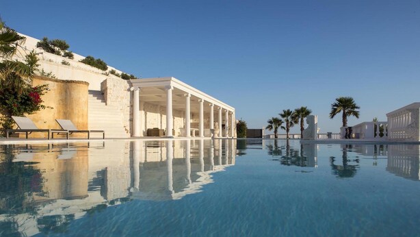 A pool with clear water reflecting a building with columns, sun loungers, and palm trees. - Aelya (Paros)