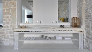 A bathroom with a stone wall, a white sink with a mirror, and a towel rack. - Aelya (Paros)