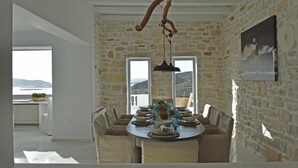 A dining area with a stone wall, a table set for six, and a view of the sea through large windows. - Aelya (Paros)