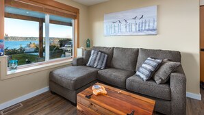 A living room with a gray sectional sofa, a wooden coffee table, and a large window offering a view of the outdoors. - Pier View Suite, Stunning Ocean View. (Campbell River)