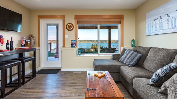 A living room with a sofa, a coffee table, a TV, and a window with a view of a lake. - Pier View Suite, Stunning Ocean View. (Campbell River)